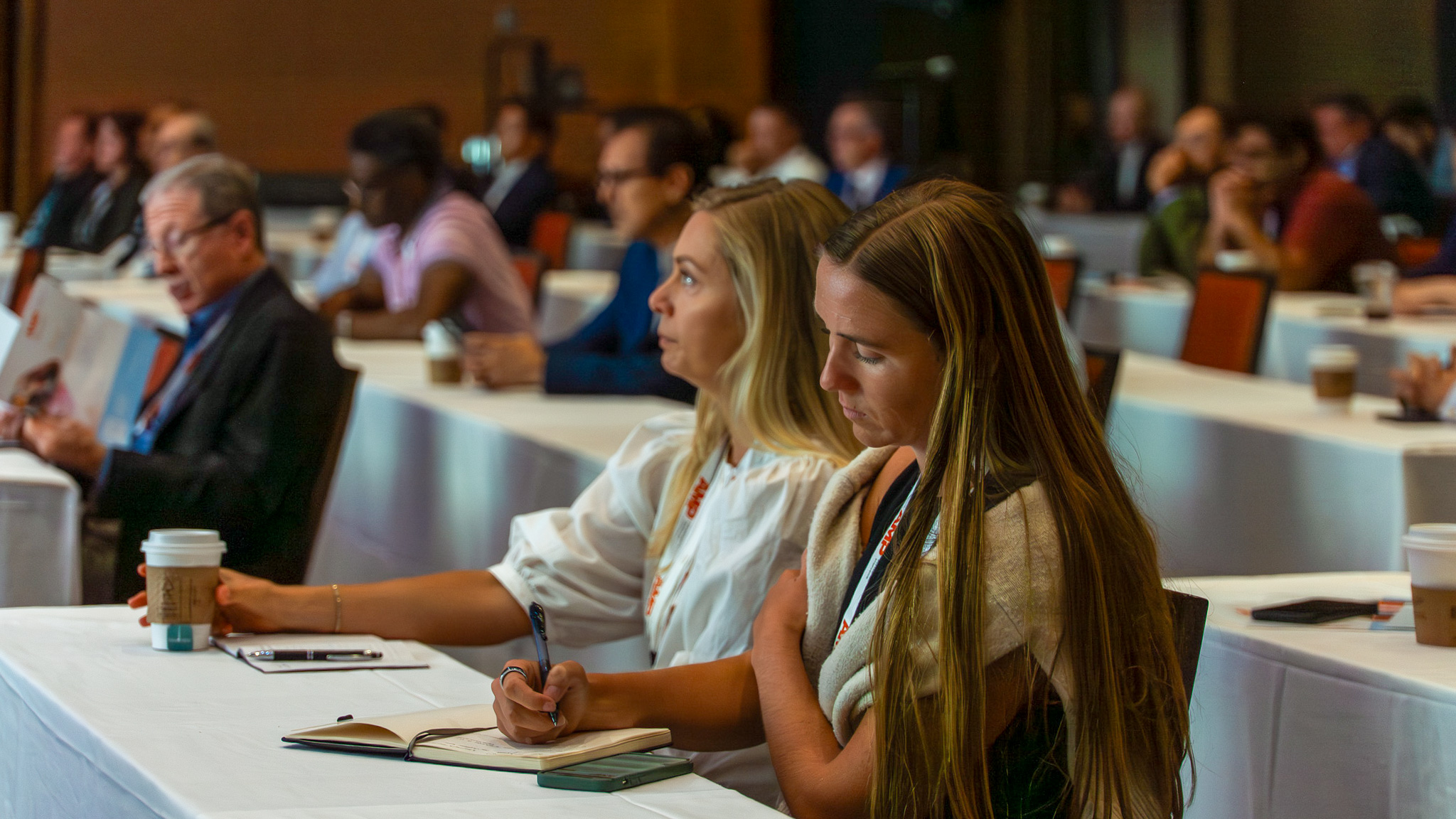Two women sit in an AMP session taking notes on the presentation.