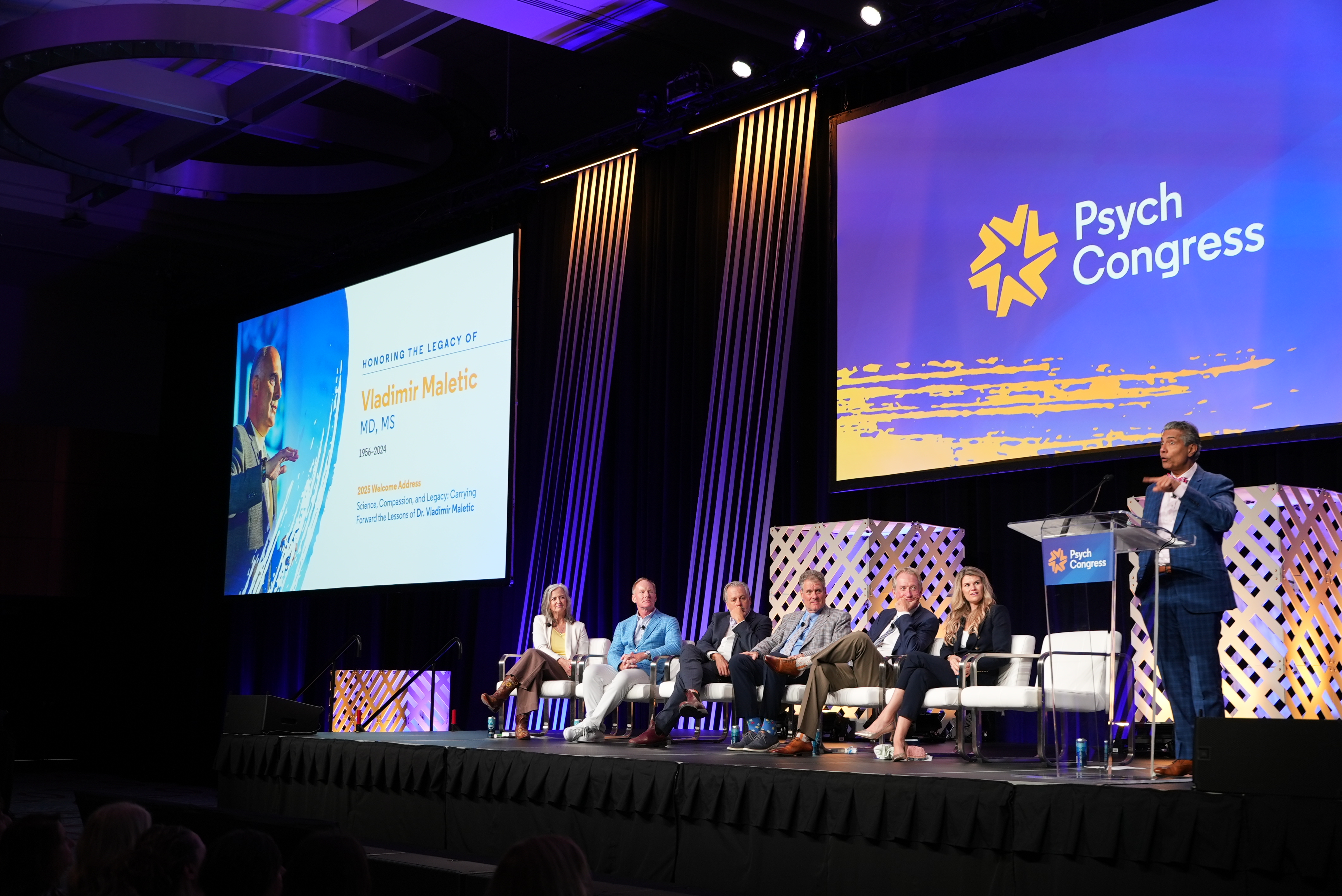 A panel of speakers look on as Rakesh Jain, MD, MPH, speaks on stage in honor of Dr Vladimir Maletic.