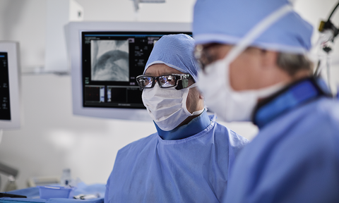 Two doctors in blue scrubs wearing white surgical masks and scrub hats. One doctor is wearing dark rimmed glasses and there is a monitor behind him.