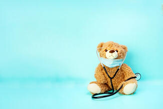 A stuffed teddy bear sits against an aquamarine background with a medical mask 