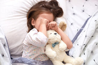 A sleepy child rubs their eyes while laying in bed with a teddy bear