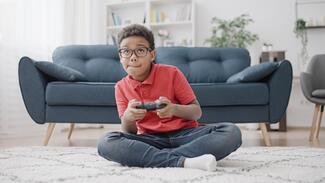 A young Black boy sits on the floor of a living room holding a video game controller and concentrated on his game