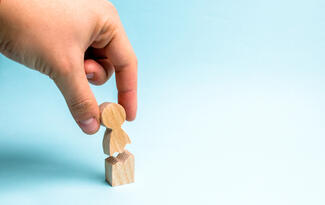 A human hand breaks off a head from a wooden figure against a light blue background