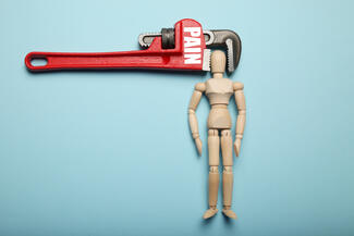 A wrench with the word 'pain' on it holds the head of a wooden figurine in a vice grip