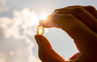 a human hand holds a Vitamin D supplement pill against a background of clouds in the sky on a sunny day