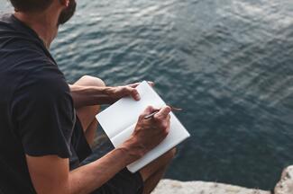 A man sits by water with a journal open on his lap for writing 