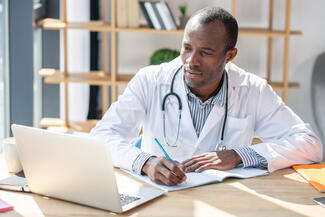 a doctor sits at a desk and takes notes while looking at a laptop