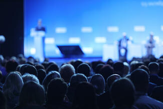 An audience at a conference listens to a speaker on stage